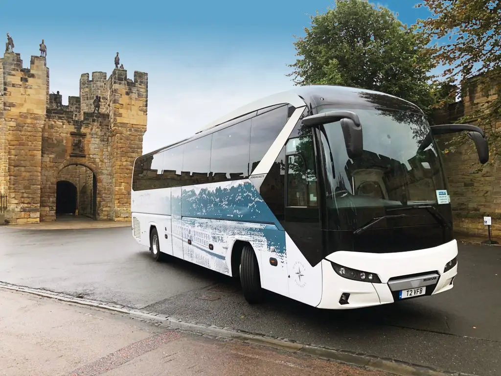 The image features a sleek and modern Harmony Voyages coach, showcasing its stylish exterior design and branding. The coach is predominantly white with elegant blue and gold accents, prominently displaying the Harmony Voyages logo. Large tinted windows offer a view of the comfortable seating inside, suggesting a luxurious travel experience. The coach is parked in a scenic location, with a backdrop of lush greenery and trees, emphasizing its suitability for exploring beautiful destinations. This image captures the essence of comfortable and sophisticated travel offered by Harmony Voyages.